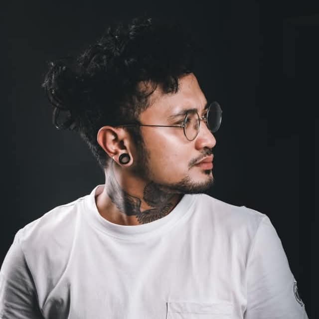 Side profile of a man with curly hair, round glasses, ear gauges, and neck tattoo.